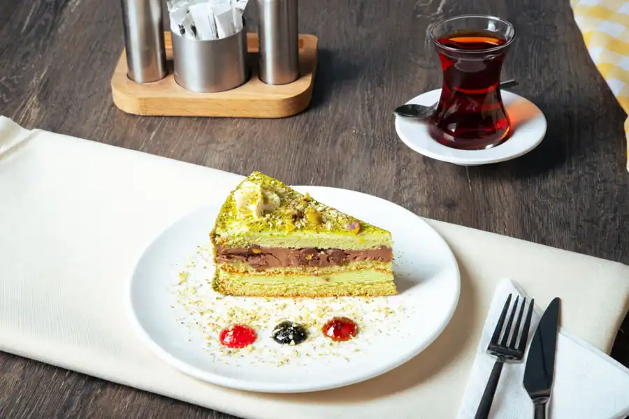 A delicious pistachio cake slice served with a glass of tea on a rustic wooden table, perfect for dessert lovers.