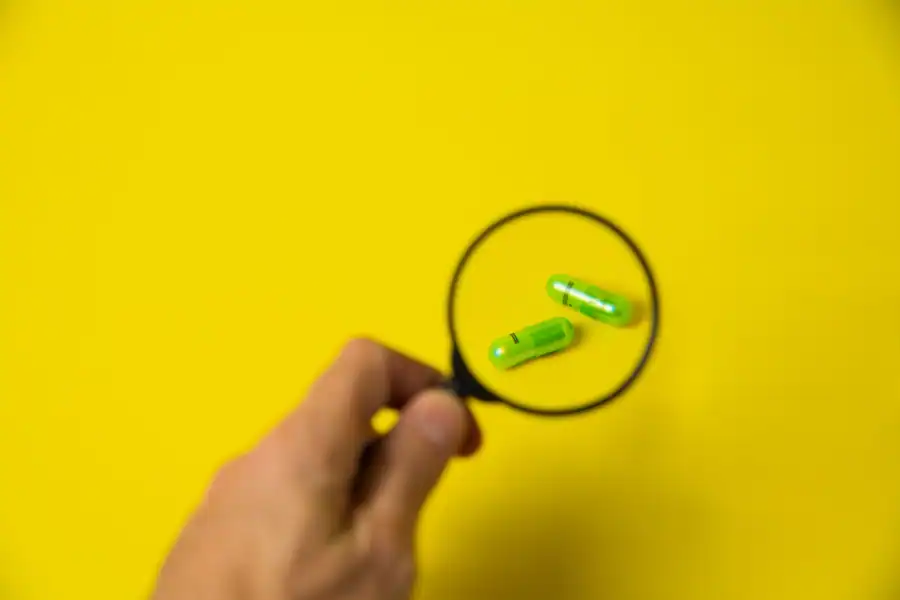 From above of crop unrecognizable male holding magnifying glass over green pills on yellow background in studio